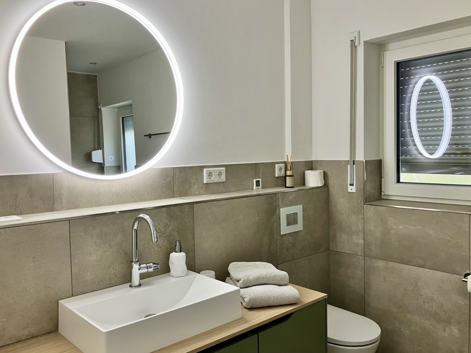 Modernes Badezimmer mit Waschtisch, beleuchtetem Rundspiegel, WC und Fenster